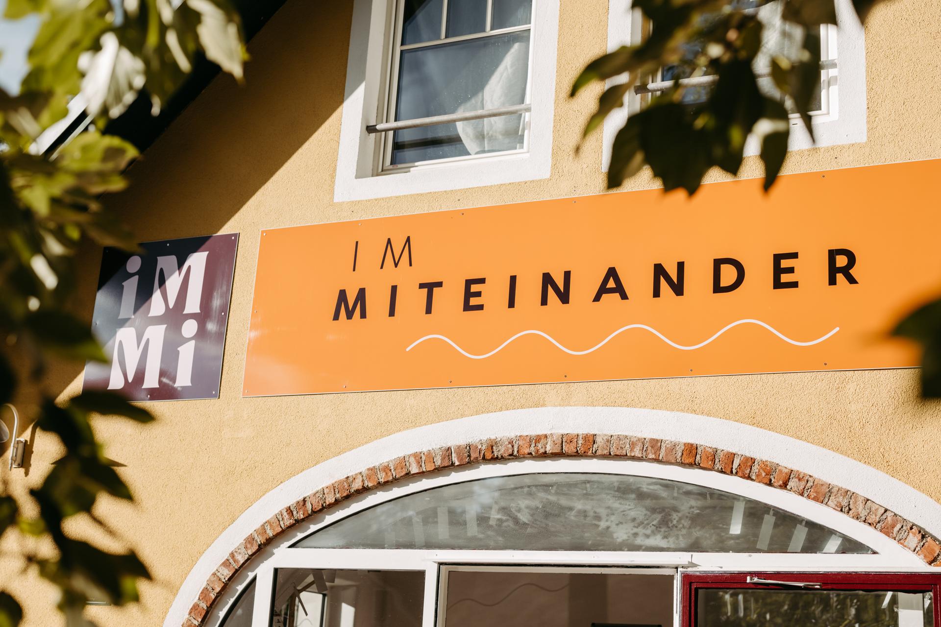 Nahaufnahme der Beschilderung am Haus: das dunkelrote iMMi-Logo neben dem orangen Schriftzug IM MITEINANDER, gerahmt von Blättern