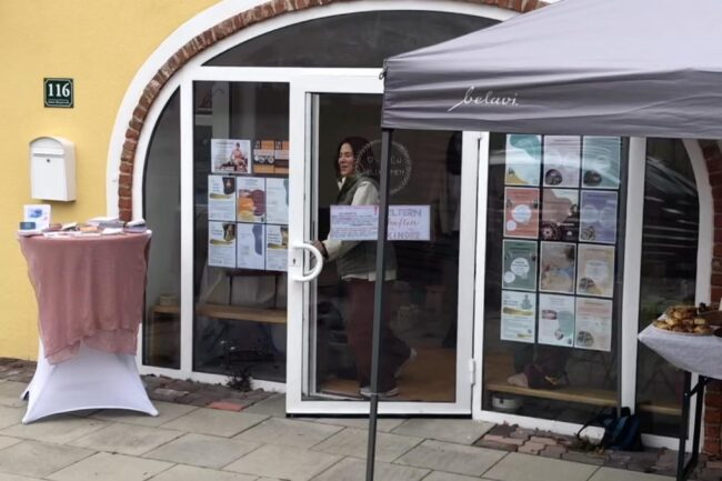 Der Eingang des IM MITEINANDER beim Come Together: ein Pavillon vor dem gelben Haus, Stehtische mit Informationsmaterial und Gebäck, im Türrahmen eine Frau
