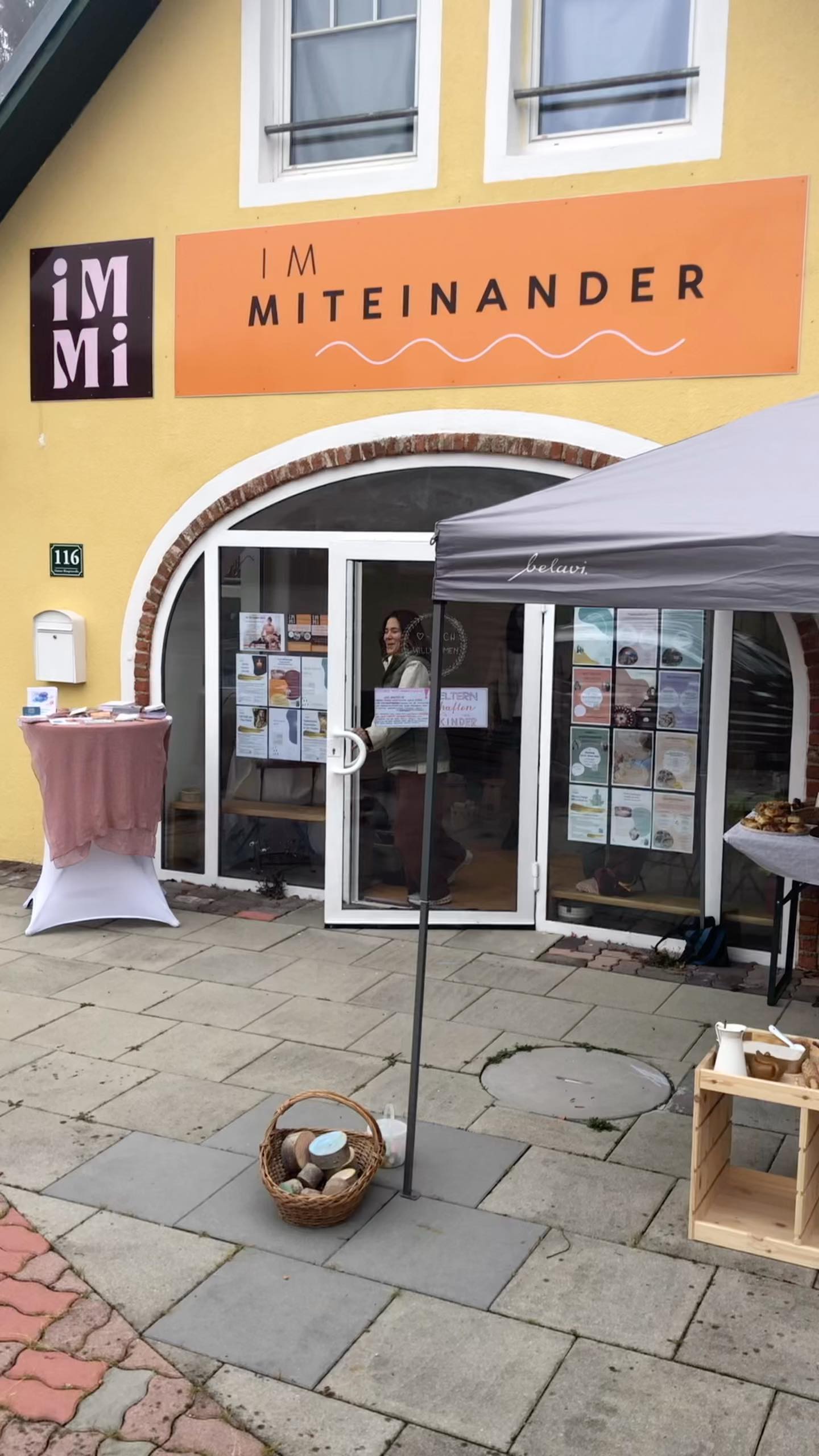 Der Eingang des IM MITEINANDER beim Come Together: ein Pavillon vor dem gelben Haus, Stehtische mit Informationsmaterial und Gebäck, im Türrahmen eine Frau