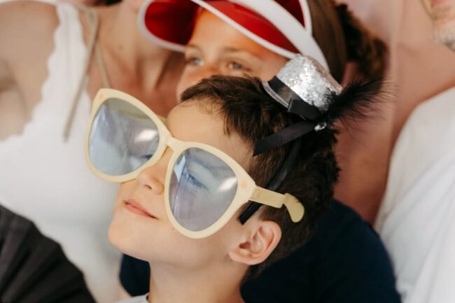 Ein Kind mit übergroßer Sonnenbrille beim Sommerfest zum einjährigen Bestehen, dahinter Erwachsene